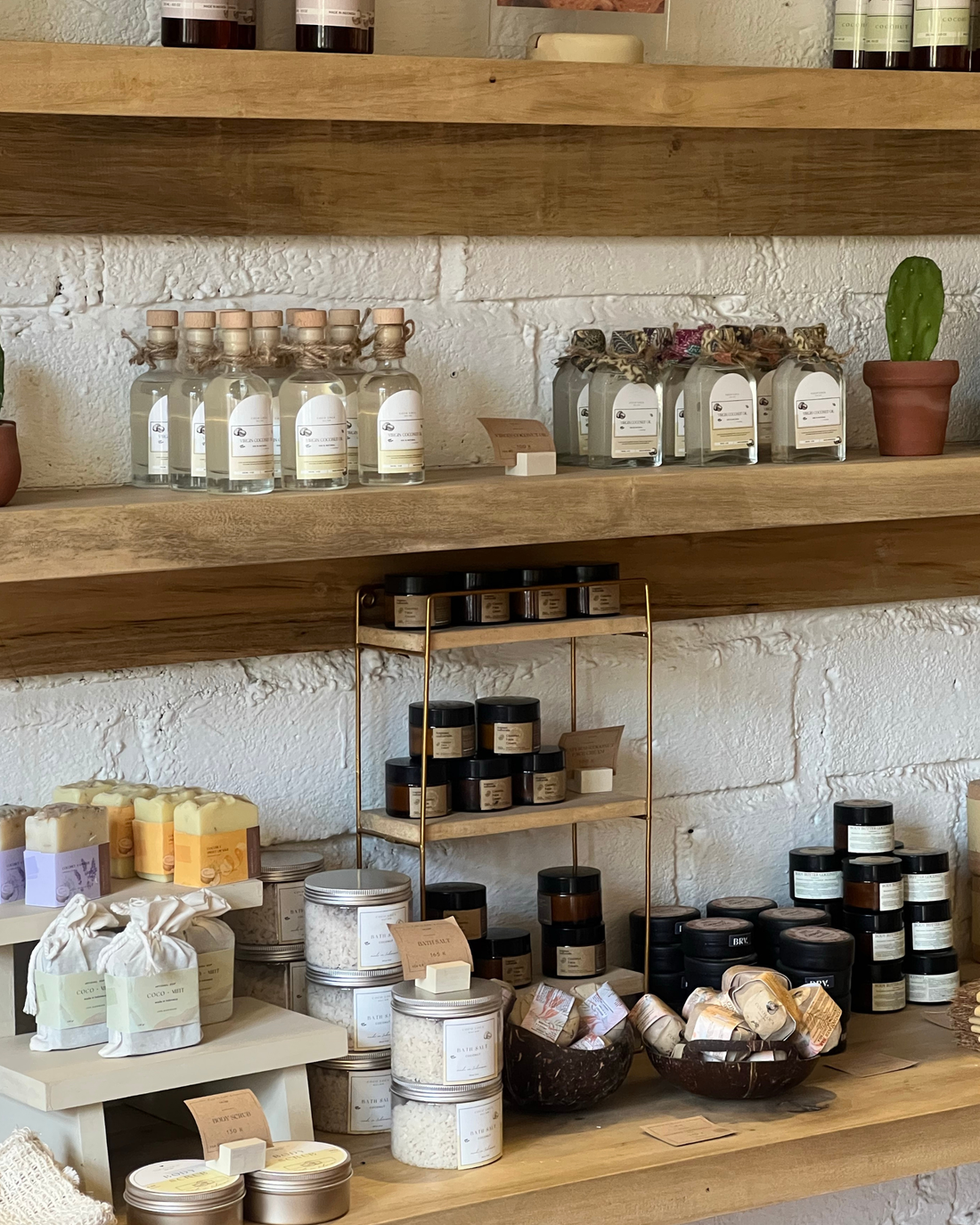 A styled retail display of artisanal home and beauty products on rustic wooden shelves against a white brick wall. The collection includes clear glass bottles with cork stoppers, amber jars, metal tins, wrapped soaps, and a small potted cactus.