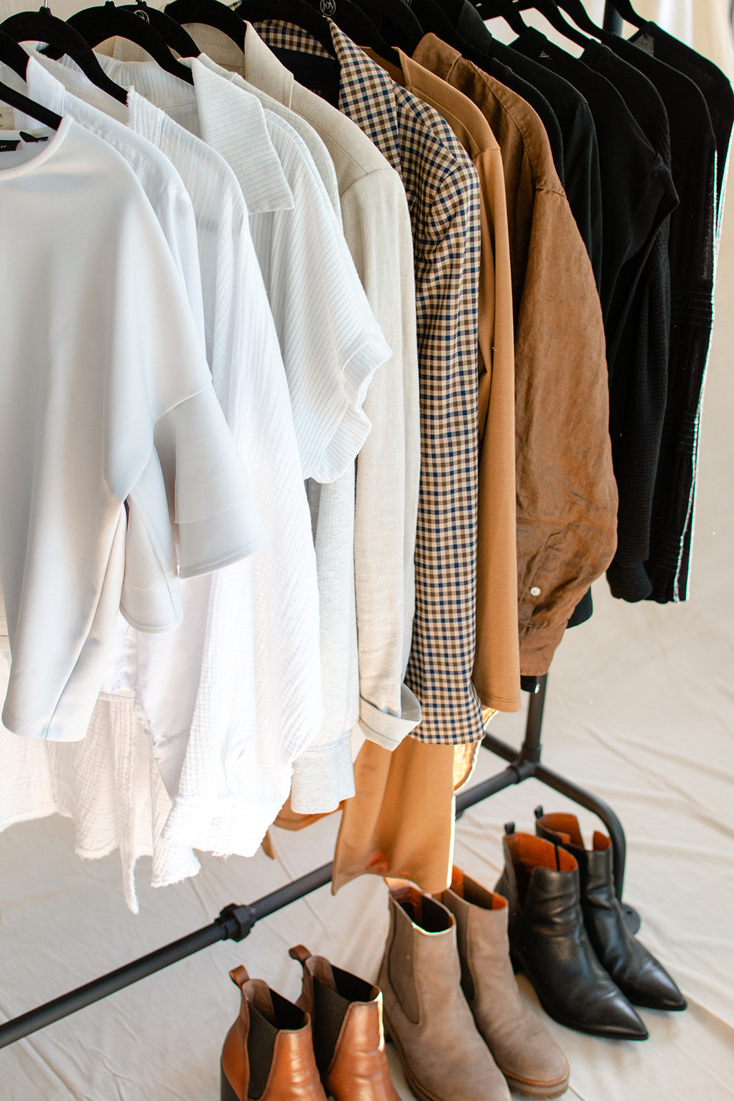A clothing rack displaying a curated collection of women's apparel, including white and beige short-sleeve tops, a brown and beige gingham shirt, tan jackets, and black tops, with matching ankle boots and Chelsea boots arranged on the floor below.